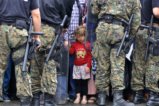 10일 마케도니아 남부에 위치한 게브젤리자에서 난민들의 국경 통과를 막아선 경비대원들을 앞에 두고 한 난민 소녀가 고개를 숙인 채 기다리고 있다. 이날 가디언 보도에 따르면 마케도니아 경비대원들이 난민 유입을 차단하기 위해 난민들에게 곤봉을 휘두르며 위협하는 모습이 동영상에 잡혀 논란이 일고 있다.