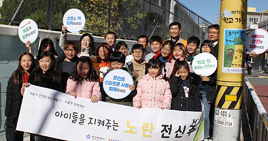 부산 사상초등학교 학생들이 스쿨존에서 감속 및 안전 운행의 필요성을 담은 노란 전신주 앞에 모여 교통안전 캠페인을 펼치고 있다. 노란 전신주 도입 후 긍정적인 반응과 효과가 확인됐다.   초록우산어린이재단 제공