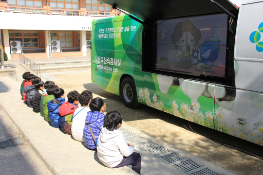 어린이들이 초등학교에 찾아온 ‘식중독 신속 검사 차량’ 앞에 나란히 앉아 멀티비전을 통해 식중독 예방 영상을 살펴보고 있다. 식품의약품안전처 제공