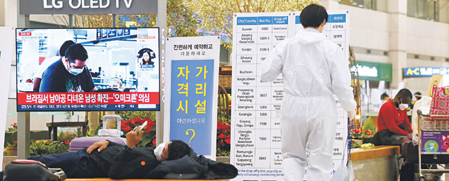 29일 오전 인천국제공항 외국인검역대기장소에서 한 입국자가 검역절차를 기다리다가 의자에 누워 휴대전화를 들여다보고 있다. 뒤편의 TV 화면에는 “브라질서 남아공 다녀온 남성 확진, ‘오미크론’ 의심” 자막이 나오고 있다.  곽성호 기자