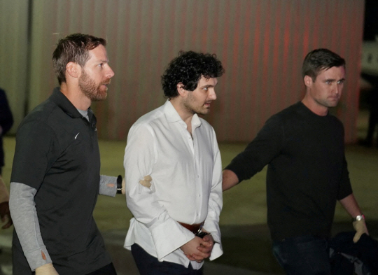 세계 3대 가상화폐 거래소였던 FTX의 창업자 샘 뱅크먼-프리드(가운데)가 21일(현지시간) 체포돼 있던 바하마의 공항에서 미국으로 송환되는 비행기를 타기 위해 수갑을 찬 채 이동하고 있다. 로이터·연합뉴스