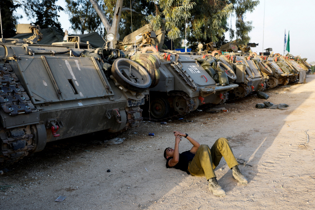 이스라엘과 하마스 간 인질 석방 및 나흘 휴전 합의가 발효되기 하루 전인 23일 한 이스라엘 군인이 이스라엘과 팔레스타인 가자지구 국경선 인근의 병력 수송 장갑차(APC) 그늘 아래 누워 휴대전화를 보고 있다. 로이터 연합뉴스
