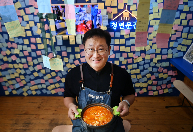 지난달 24일 ‘청년밥상문간’ 이대점에서 만난 이문수 신부. 푸짐한 1인분 김치찌개 냄비를 든 이 신부가 손님들이 붙여 둔 메모지들을 배경으로 웃고 있다. “맛있다”는 감사 인사뿐만 아니라 취업 등 개인의 소망을 쓴 경우도 있다. 윤성호 기자